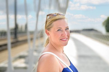 Dost canlısı, orta yaşlı sarışın bir kadın yaz güneşinde bir köprüde yakın plan bir portrede kameraya bakıp gülümsüyor.