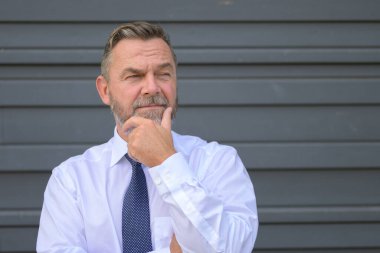 Entrikacı işadamı ciddi bir düşünceli ifadeyle yan tarafa bakıyor ve eli gri bir dış duvarın önünde dikilirken çenesini kaldırıyor.