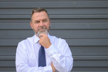 Eli çenesinde gri bir dış duvarın önünde dikilirken düşünceli işadamı derin derin bir tefekkür içinde yan tarafa bakıyor.