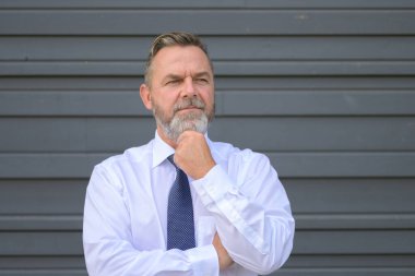 Eli çenesinde gri bir dış duvarın önünde dikilirken düşünceli işadamı derin derin bir tefekkür içinde yan tarafa bakıyor.