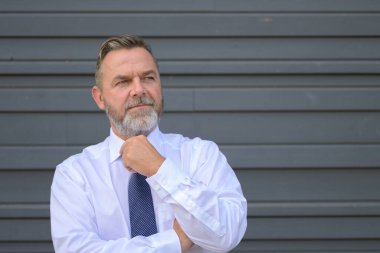 Entrikacı işadamı ciddi bir düşünceli ifadeyle yan tarafa bakıyor ve eli gri bir dış duvarın önünde dikilirken çenesini kaldırıyor.