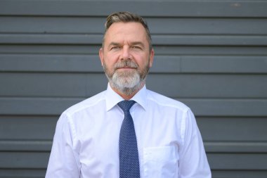 Gri bir dış duvarın önünde duran esrarengiz sakallı işadamı ön ve omuz portresindeki düşünceli bir ifadeyle kameraya sessizce bakıyor.