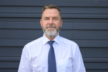 Gri bir duvarın önünde poz veren kıdemli işadamı ön ve omuz portresindeki sessiz bakışlarla kameraya dikkatlice bakıyor.