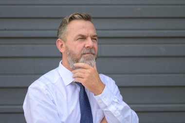 Endişeli iş adamı, eli çenesinde derin bir tefekküre dalmış ve ciddi bir şekilde dış duvarın önünde duruyor.