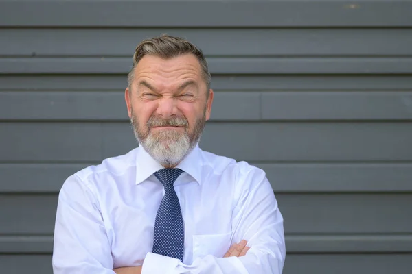 Businessman pulling a quirky face screwing up his eyes and squinting at the camera as he poses with crossed arms in front of a grey exterior wall