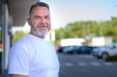 Sakallı, kendine güvenen atletik bir adam sessiz bir sokakta dikiliyor ve özenli bir şekilde kameraya bakıyor.