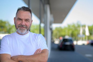 Rahatlamış, kendine güvenen, tişörtlü yaşlı bir adam sokakta dostça bir gülümsemeyle kameraya bakıyor.