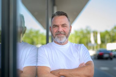 Rahatlamış, kendine güvenen, tişörtlü, büyük bir pencereye yaslanmış, caddede camdan yansıyan, dostça bir gülümsemeyle kameraya bakan yaşlı adam.