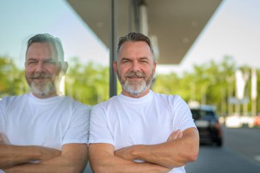 Rahatlamış, kendine güvenen, tişörtlü, büyük bir pencereye yaslanmış, caddede camdan yansıyan, dostça bir gülümsemeyle kameraya bakan yaşlı adam.