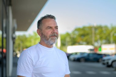 Düşünceli sakallı son sınıf öğrencisi açık hava otoparkında dikilip kafa ve omuz portresindeki düşünceli ifadesiyle yan yana bakıyor.