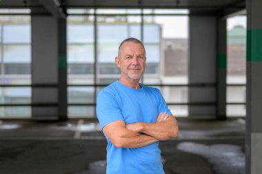 Gündelik mavi tişört giyen olgun, kendine güvenen bir adam modern bir şehir otoparkında kollarını kavuşturmuş duruyor..