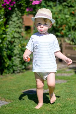 Çizgili tişört ve hasır şapkalı genç bir çocuk elinde küçük kırmızı bir domates tutarken çimenlerde yalınayak yürüyor..