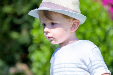 Çizgili tişört ve hasır şapkalı genç bir çocuk açık havada güneş ışığında dikilirken düşünceli görünüyor..