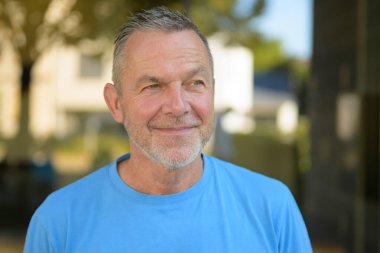 Mavi gömlekli, kendine güvenen olgun bir adam güneşli bir günde, arka planı bulanık ve yumuşak bir şekilde dışarıda dururken nazikçe gülümsüyor..