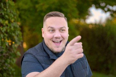 Heyecanlı bir ifadeye sahip mutlu bir genç adam arka planda yeşil ağaçlarla dışarıda dikilirken ileri doğru işaret eder ve güler..