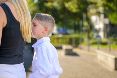 Beyaz tişörtlü genç, sevgiyle annesine doğru eğiliyor. Açık havada, samimi bir aile ortamında sevgi ve sıcaklık gösteriyor..