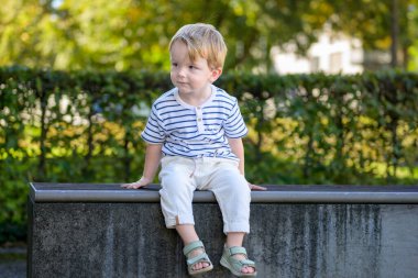Dışarıda alçak bir duvarda oturan çizgili gömlek ve açık renk pantolon giyen sevimli genç bir çocuk, sıcak gün ışığında meraklı ve rahat görünüyor..