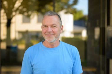 Dost canlısı, orta yaşlı, kısa gri saçlı, sakallı açık mavi gömlekli, güneşli bir günde dışarıda beklerken sıcak bir şekilde gülümseyen bir adam..