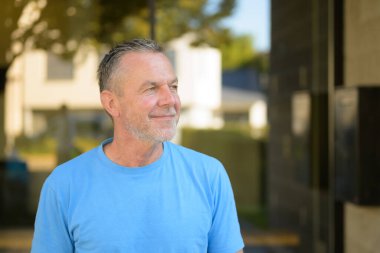 Kısa gri saçlı ve sakallı, kendine güvenen, açık mavi gömlekli, güneşli bir günde dışarı bakarken nazikçe gülümseyen olgun bir adam..