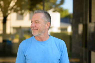 Kendine güvenen, kısa gri saçlı, sakallı, mavi tişörtlü, gün ışığında dışarı bakarken nazikçe gülümseyen olgun bir adam..