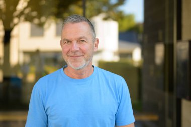 Sakin ve kendinden emin, kısa gri saçlı, sakallı açık mavi tişörtlü, güpegündüz dışarıda dikilen, sakin ve kendinden emin bir adam..