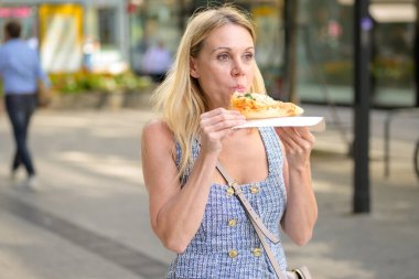 Sarışın kadın güneşli bir günde şehirde yürürken taze pizza yiyor..