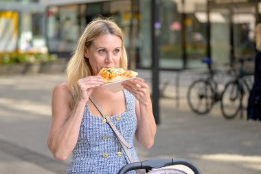 Sarışın kadın güneşli bir günde bebek arabasıyla gezerken dışarıda pizza yiyor..