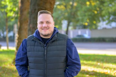 Güz boyunca güneşli yeşil bir parkta kapüşon ve yelek giyen kendine güvenen genç bir adam..