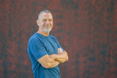 Kısa saçlı, sakallı, mavi tişörtlü, kırmızı desenli duvarda kolları çaprazlanmış, kendinden emin orta yaşlı bir adam..