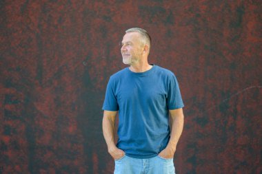 Kısa saçlı, sakallı, mavi tişörtlü, kot pantolonlu, açık havada kırmızı bir duvara bakan, kendinden emin orta yaşlı bir adam..