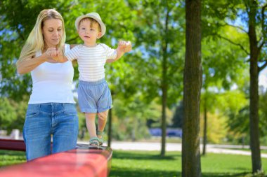 Gülümseyen bir anne, güneşli yeşil bir parkta kırmızı denge demetinde yürüyen şapkalı ve çizgili gömlekli oğluna yardım eder..