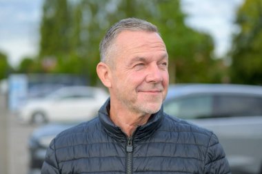 Koyu renk ceketli olgun bir adamın portresi Parlak bir günde dışarı bakarken hafifçe gülümsüyor.