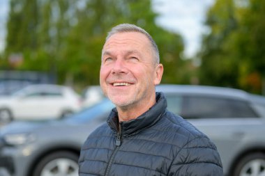 Koyu renk ceketli neşeli olgun bir adamın portresi serin bir günde dışarıda dikilirken neşeli bir gülümsemeyle yukarı bakıyor..