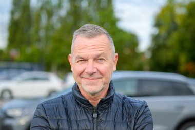 Koyu renk ceketli neşeli, olgun bir adamın portresi serin bir günde dışarıda dikilirken neşeyle göz kırpıyor..