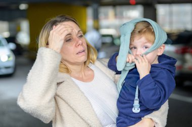 Bitkin bir anne, hasta bebeğini modern bir otoparkta taşıyarak günlük ebeveynlik süresince stres ve endişe gösteriyor..