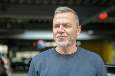 Kısa gri saçlı, sakallı, kendine güvenen olgun bir adam. Kapalı yerde, bulanık şehir geçmişine karşı duruyor..