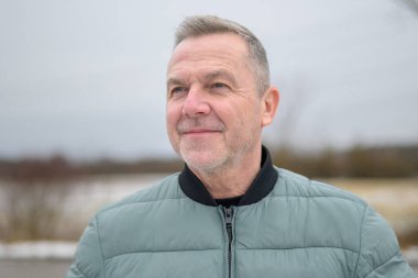 Yeşil ceketli, gülümseyen olgun bir adam bulutlu bir kış gününde açık havaya bakıyor..