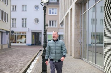 Gülümseyen olgun bir adam modern bir şehir caddesinde yürüyor, güveni, yaşam tarzını, bağımsızlığı ve aktif yaşlılığı temsil ediyor..