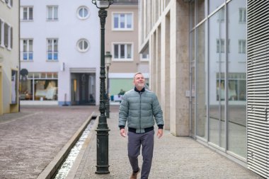 Olgun bir adam sakin bir şehir caddesinde, mimari, asfalt ve doğal gün ışığıyla sakin bir anın tadını çıkararak güvenle yürüyor..