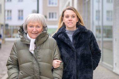 Bir şehir sokağında yetişkin kızıyla kol kola yürüyen bir anne. Aile desteği, bakım, güven ve güçlü bir çoklu-kuşak ilişkisini temsil ediyor..