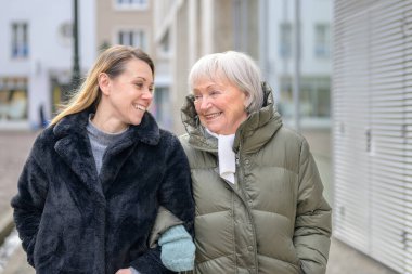 Modern bir şehirde yetişkin kızıyla birlikte yürüyen mutlu yaşlı kadın, iki nesil arasındaki sıcaklığı, güveni ve duygusal bağı paylaşıyor..