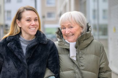 Modern bir şehirde, yetişkin kızına yakın duran gülümseyen yaşlı kadın nesiller arasındaki sevgi, güven, sıcaklık ve duygusal bağı gösteriyor..
