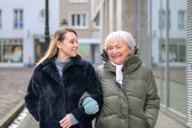 Farklı nesillerden iki kadın modern bir şehir ortamında birlikte gülümsüyor, aile bağlarını, sıcaklığı, güveni ve pozitif yaşlanmayı gösteriyorlar..