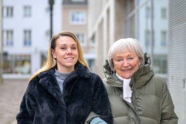 Farklı nesillerden iki kadın modern bir şehir ortamında birlikte gülümsüyor, aile bağlarını, sıcaklığı, güveni ve pozitif yaşlanmayı gösteriyorlar..