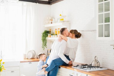 Genç ebeveynler modern mutfağın arka planında sarılıp öpüşürler. Küçük bir oğul onlara bakar ve babasına sarılır..