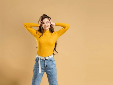 Turuncu kazaklı, uzun kıvırcık saçlı genç ve çekici bir kız ellerini pastel turuncu stüdyo arka planında tutuyor..