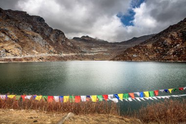 Himalayalar 'ın ortasındaki Sikkim, Hindistan' daki Changu Gölü manzarası. Görüntü, önplanda dalgalanan bir dizi geleneksel renkli Budist dua bayraklarını, durgun suyu ve dramatik bulutlu bir gökyüzünün altındaki sarp, kayalık dağ yamaçlarını çerçevelemektedir..