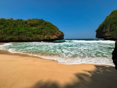 Okyanus dalgalarının açık mavi gökyüzünün altındaki iki yemyeşil kıyı uçurumu arasında altın kumda yuvarlandığı güzel tropik plaj manzarası. Huzurlu deniz manzarası seyahat, tatil ve doğa arka plan kavramları için idealdir..