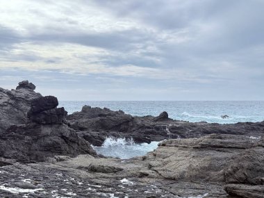 Okyanus dalgalarının dramatik bulutlu bir gökyüzü altında kıyıya vurduğu manzaralı kayalık kıyı manzarası. Seyahat, doğa ve çevresel temalar için uygun doğal doğa manzarası.