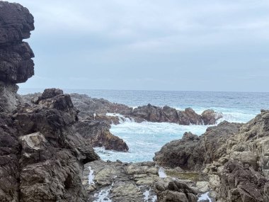 Okyanus dalgalarının bulutlu bir gökyüzünün altındaki kayalara çarptığı Rocky kıyı manzarası. Seyahat, doğa ve çevre temaları için uygun doğal deniz manzarası.